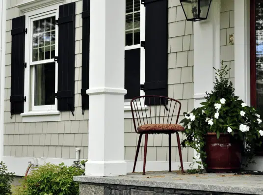 Front porch of a modern home featuring a cozy metal chair and a vibrant flower pot with white blooms, complemented by black shutters on the windows and a welcoming atmosphere.