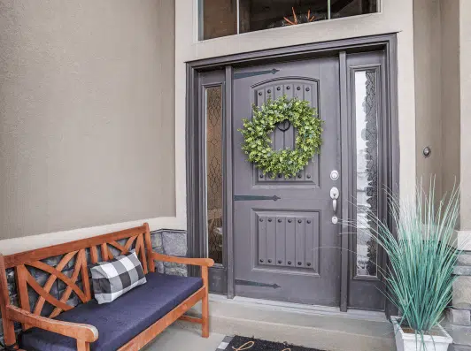 Front porch featuring a stylish dark door adorned with a green wreath, accompanied by a wooden bench with a checkered cushion and decorative plants.