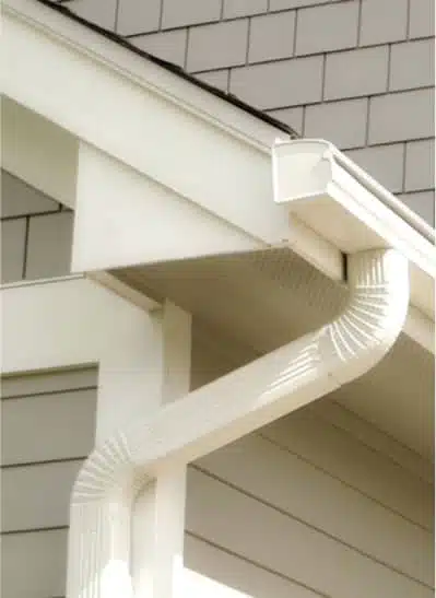 Close-up of a white gutter system installed on a house, showcasing the downspout and eavestrough against a light-colored shingle exterior. Ideal for illustrating home drainage solutions and roofing details.