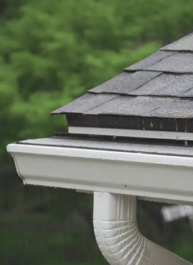 Close-up of a roof gutter system with shingles, capturing rainwater during a downpour, surrounded by lush greenery in the background.