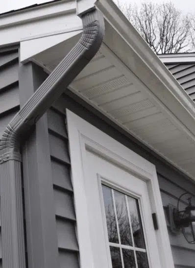 Close-up view of a home's exterior featuring a gray vinyl siding, white trim, a door with multiple panes, and a downspout connected to the gutter system, illustrating effective rainwater management.