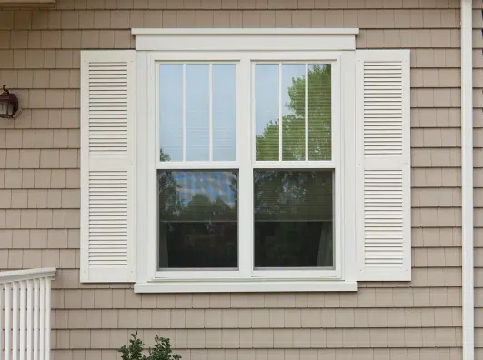 White window with shutters on a beige house exterior, featuring four panes of glass and a view of greenery reflected in the window.